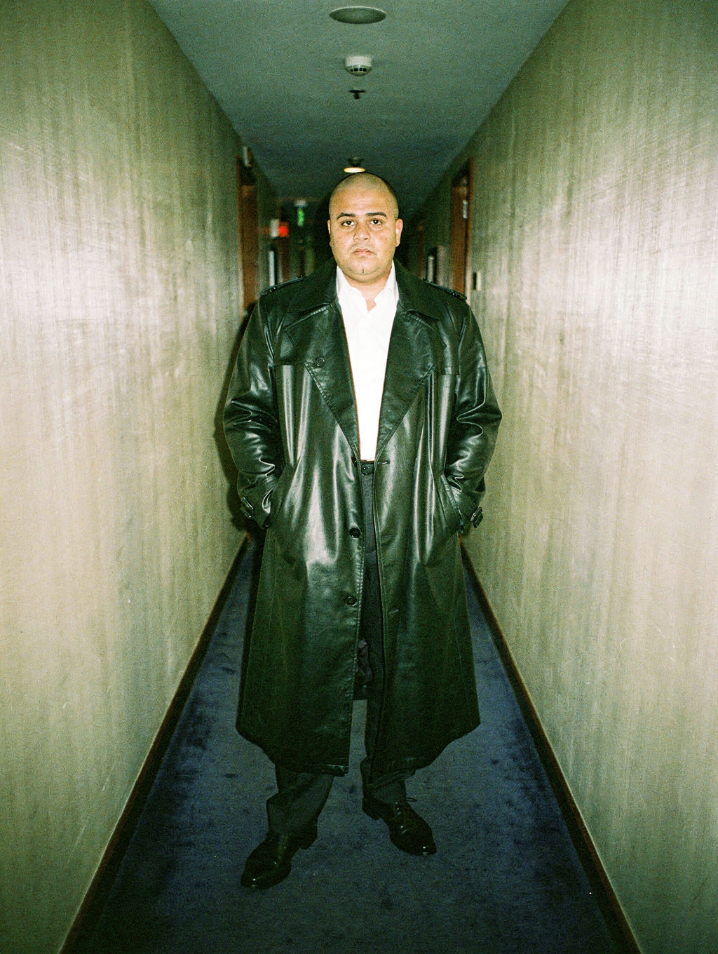 Man in a black leather trench coat in a flash-lit hotel corridor