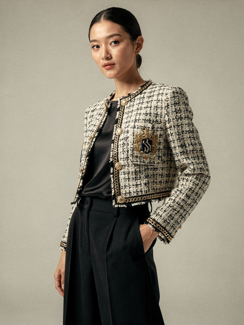 Model in a cropped bouclé jacket with an embroidered crest and gold chain trim